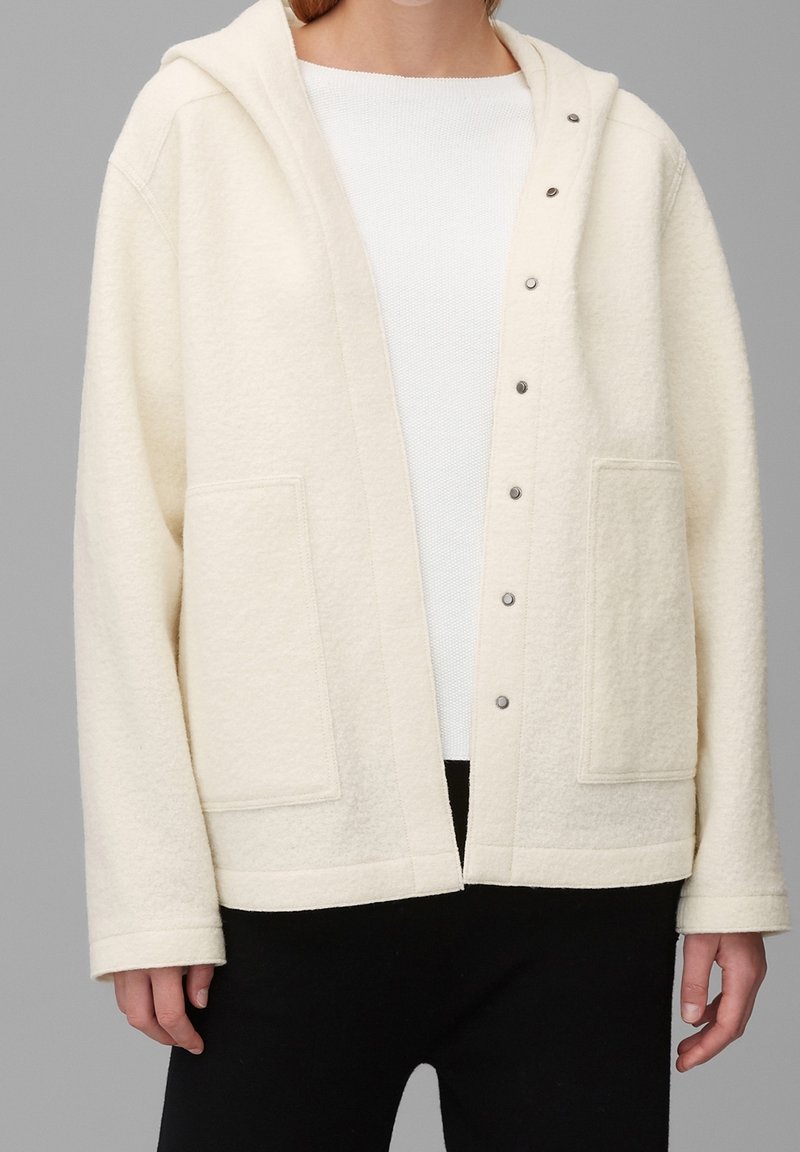 Personne portant une veste à capuche écrue avec des boutons-pression et de grandes poches avant sur un haut blanc et un pantalon noir.