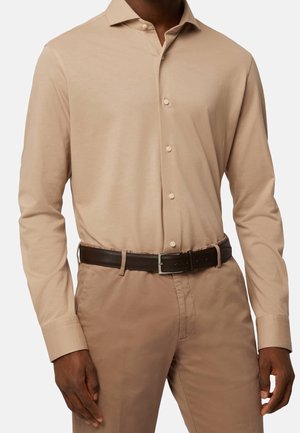 Muž nosí béžovou košili s dlouhým rukávem, zapnutou v světle hnědých kalhotách s tmavě hnědým koženým pásem.