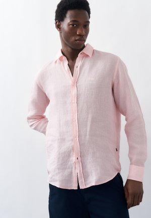 Mann trägt ein hellrosa langärmliges Hemd mit Knöpfen und dunkle Hose, steht mit einer Hand hinter dem Rücken vor einem schlichten Hintergrund.