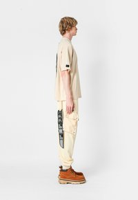 Camiseta oversized beige con diseño gráfico, combinada con pantalones cargo crema que incluyen bolsillos y detalles en texto negro, y botas marrones con cordones.