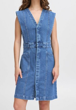 Niebieska, bezrękawnikowa sukienka dżinsowa z głębokim dekoltem w kształcie litery V, przednim szwem, pętlami na pasek w talii i bocznymi kieszeniami, noszona przez stojącą osobę.