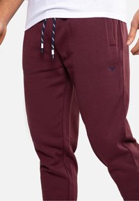 Burgundské tepláky vyrobené z měkké látky s elastickým pasem, námořnickými šňůrkami a malým logem. Obsahují boční kapsy a zúžené nohavice.