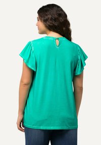 Top in cotone verde con maniche arricciate, dotato di un dettaglio a chiave sul collo e una vestibilità comoda. Sfondo neutro.