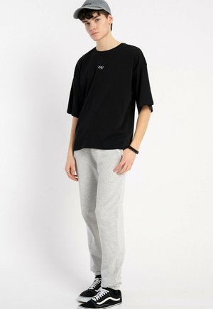 Czarna, oversize'owa koszulka z małym logo, szare dresy i czarne sneakersy z białymi akcentami. Model nosi szary czapkę.
