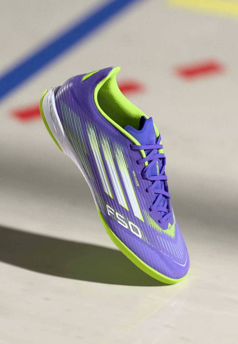Scarpa sportiva viola con superficie testurizzata, caratterizzata da accenti verdi, strisce bianche e un design a profilo basso. I lacci sono viola.