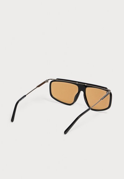 Lunettes de soleil à monture noire avec des verres rectangulaires teintés ambre et des branches métalliques fines posées sur une surface blanche.
