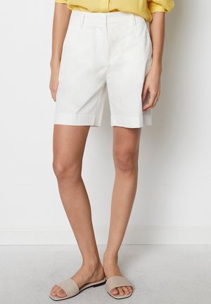 Frau mit weißen, maßgeschneiderten knielangen Shorts, gelbem Hemd und beigen Sandalen, die vor einer schlichten weißen Wand steht.