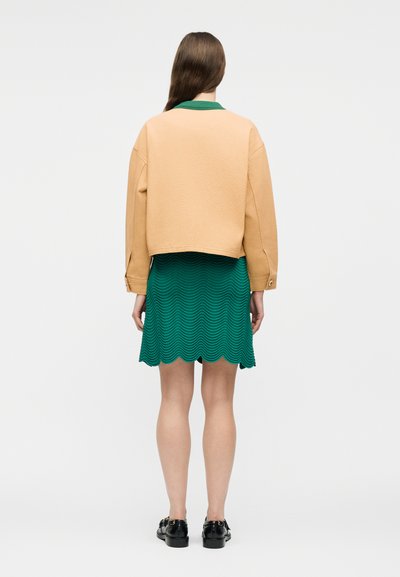 Γυναίκα που στέκεται δίνοντας την πλάτη της, φοράει μπεζ κοντό σακάκι, πράσινη υφή φούστα με κυματιστό τελείωμα και μαύρα μοκασίνια, μπροστά σε απλό φόντο.