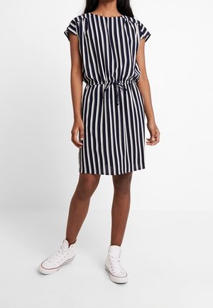 Femme portant une robe rayée verticale bleue et blanche jusqu'aux genoux avec une taille nouée et des baskets montantes blanches, se tenant devant un fond blanc.