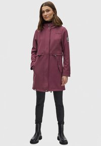 Maroon waterproof coat with a high collar, zip fastenings, side pockets, adjustable waist, and ribbed cuffs, paired with black trousers and boots.