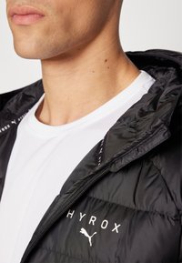 Juodas paminkštintas džemperis su užtrauktuku priekyje, ant kurio yra prekės ženklo "HYROX" ir "Puma" logotipas. Dėvimas ant baltos tekstūruotos marškinėlių.