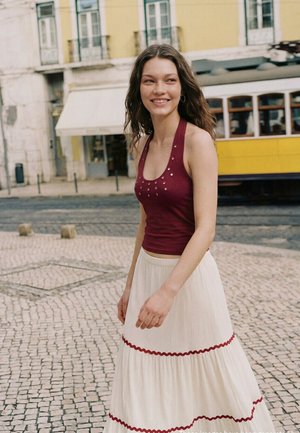 Jeune femme aux cheveux ondulés portant un haut sans manches bordeaux et une jupe blanche, marchant dans une rue pavée avec un tramway jaune en arrière-plan.