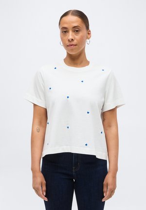 Hölgy hátrafésült hajjal, fehér rövid ujjú ingben, amelyen apró kék szívminták vannak, és sötét farmernadrágot visel, sima háttér előtt áll.