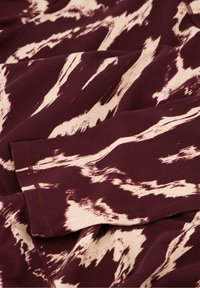 Burgunderfarbener Stoff mit einem cremefarbenen abstrakten Muster, der eine glatte Textur und einen weichen Fall aufweist. Mit kontrastierendem Stich entlang der Kanten.