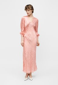 Femeie îmbrăcată într-o rochie lungă, mătăsoasă, roz cu flori și mâneci bufante, stând pe un fundal simplu, deschis la culoare, purtând pantofi albi cu vârf ascuțit.