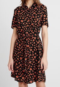 Kvinna klädd i en knälång svart klänning med röd och orange leopardmönster, korta ärmar, krage, knäppt fram och med ett knytbart midjebälte.