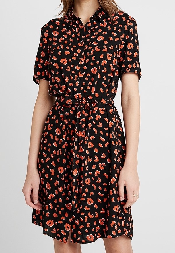 Kvinna klädd i en knälång svart klänning med röd och orange leopardmönster, korta ärmar, krage, knäppt fram och med ett knytbart midjebälte.