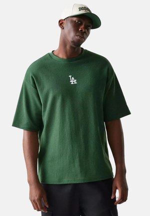 Hombre con camiseta verde con logo de LA y gorra blanca de los Dodgers, de pie contra un fondo claro y liso.