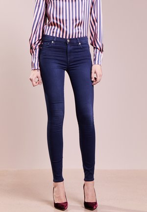 Femme portant un jean skinny bleu foncé, un chemisier à rayures verticales roses et bleues, et des chaussures à talons hauts bordeaux, debout sur un sol beige.