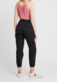 Pantalons noirs fuselés avec des poignets élastiques, dotés de deux poches arrière. Porté avec un haut floral rose et des baskets blanches avec des accents bordeaux.