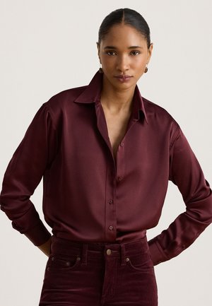 Vrouw met donker haar in een lage knot, draag een diep bordeaux satijnen blouse en bijpassende corduroy broek tegen een effen lichte achtergrond.