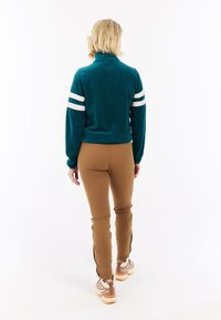 Pullover in pile turchese con colletto alto e righe bianche sulle maniche, abbinato a pantaloni marroni aderenti e sneakers chiare.