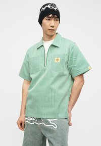 Νεαρός άντρας που φοράει μαύρο σκουφί, κοντομάνικο πουκάμισο με κάθετες πράσινες και λευκές ρίγες και φερμουάρ, και ανοιχτό μπλε τζιν με λευκά σχέδια.