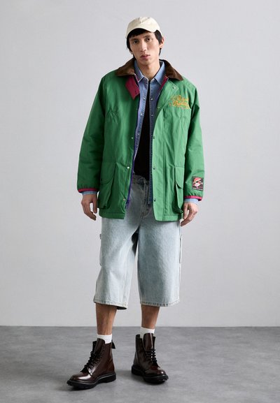 Grøn yderjakke med brun krave, multikortdesign, båret over en blå skjorte og sort top. Lyseblå brede shorts og brune snørebåndsstøvler.