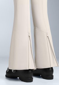 Beżowe spodnie rozszerzane z bocznymi zamkami i gładką teksturą, połączone z czarnymi butami z metalowymi akcentami i zaokrąglonym obcasem.
