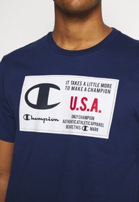 Muž v námořnickém modrém tričku s bílou nášivkou, na které je napsáno: "Chce to něco navíc, abychom se stali šampiony USA. Autentické sportovní oblečení."