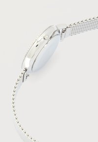 Ασημένιο ρολόι χειρός με μεταλλικό πλεκτό λουράκι, που παρουσιάζει το πλάι και το κουμπί ρύθμισης σε ανοιχτό φόντο.