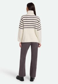 Pullover di maglia grossa color crema con strisce orizzontali grigie scure, dotato di un alto colletto e di un orlo a coste. Abbinato a pantaloni scuri e scarpe nere.