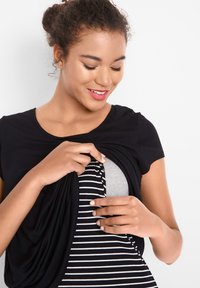 Haut noir à manches courtes, avec un design drapé. En dessous, un vêtement rayé gris et blanc est partiellement visible.