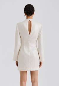 Krátke biele šaty s flitrami, dlhými rukávmi a hlbokým výrezom na chrbte, s elegantným siluetom a textúrovaným povrchom.