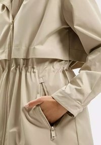 Beżowa kurtka wodoodporna z zamkiem z przodu, elastycznym pasem oraz bocznymi kieszeniami. Gładka faktura, luźny krój i długie rękawy.