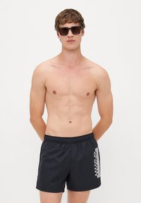 Shorts de bain noirs avec taille élastique, arborant un logo imprimé en blanc sur un côté. Modèle portant des lunettes de soleil, sans t-shirt.