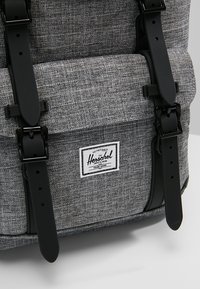 Poche de sac à dos en tissu gris texturé avec des bretelles réglables noires et une étiquette de marque Herschel blanche au centre avant.