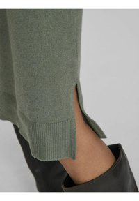 Green knit sweater with ribbed hem, featuring side slits. Black leather boot partially visible at the bottom.