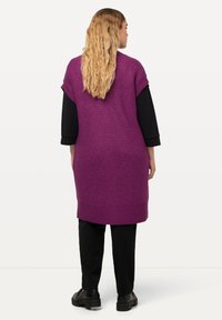 Lila stickad tunika, ärmlös med en ribbad textur, lagd över en svart långärmad topp, ihop med svarta byxor och kraftiga skor.