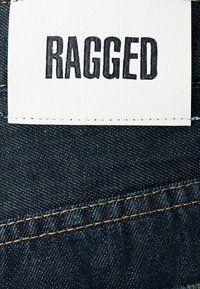 Denim tkanina s bijelom kožnom etiketom urezanom s natpisom "RAGGED". Etiketa je prišivna na tamnoplavi denim, s zlatnim koncem.