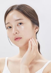 Porträtt av en person med slät hy, naturlig makeup och långt mörkt hår. Deras hand vilar på ansiktet, med en neutral bakgrund.