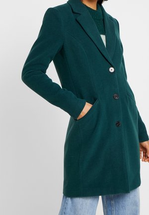 Kvinde iført en mørkegrøn lang skræddersyet frakke med tre knapper og hænderne i sidelommerne, kombineret med lyseblå jeans.