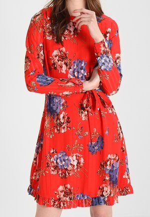 Frau trägt ein langärmeliges rotes Kleid mit blauen und pinken Blumenmustern, einem Taillenband und einem Rüschen-saum, steht mit verschränkten Armen.