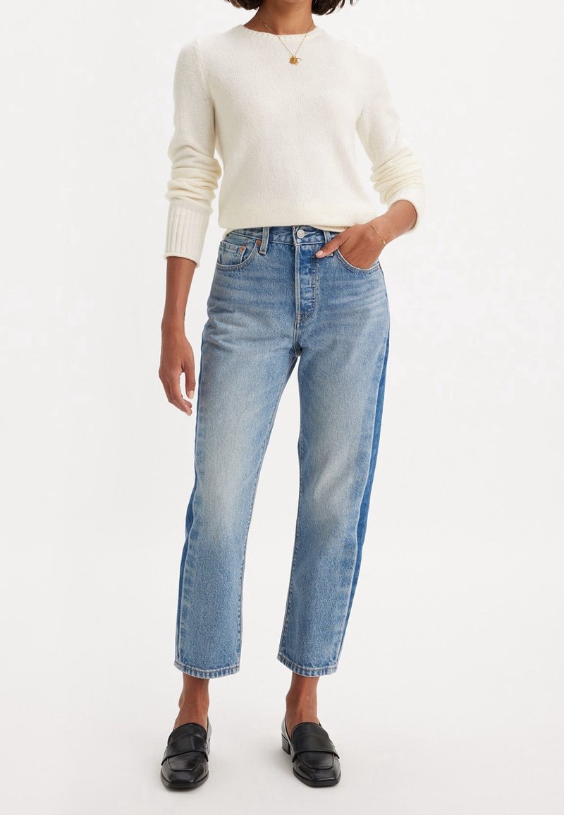 Pull crème clair, jeans bleu à jambe droite et mocassins noirs. Le jean est taille haute avec une finition délavée, orné de détails métalliques discrets.