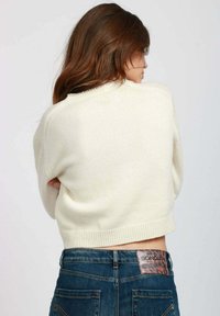 Maglione bianco a maglia con design cropped e bordo a coste, abbinato a jeans denim blu con una patch in pelle visibile in vita.