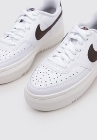 Zapatillas de cuero blanco con un acabado texturizado, que presentan una suela blanca, detalles perforados en la punta y un logo de swoosh de Nike en marrón.