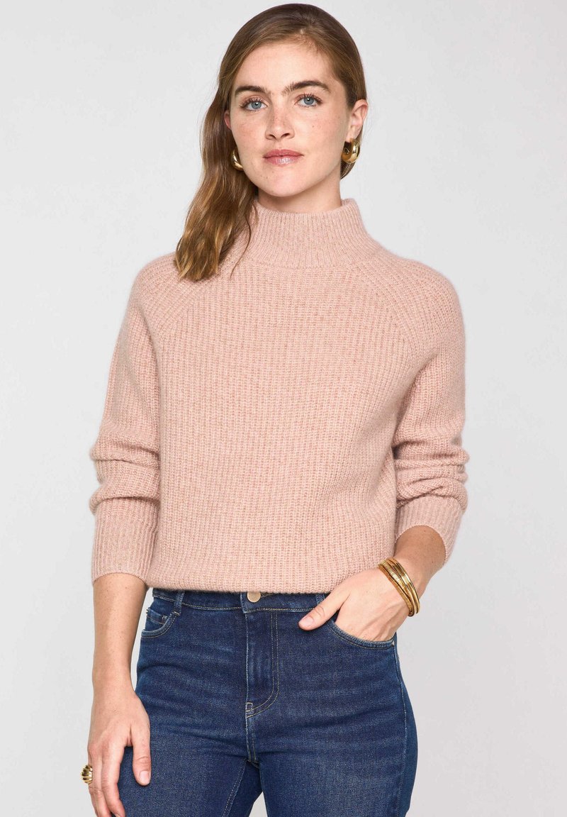 Une femme aux cheveux ondulés porte des boucles d'oreilles en or, un pull à col roulé côtelé rose, un jean bleu foncé, des bracelets en or et une bague.