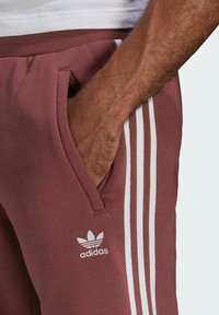 Bordové sportovní kalhoty s bílým designem se třemi pruhy a logem. Vyrobené z měkkého materiálu, obsahují boční kapsy a elastický pas.