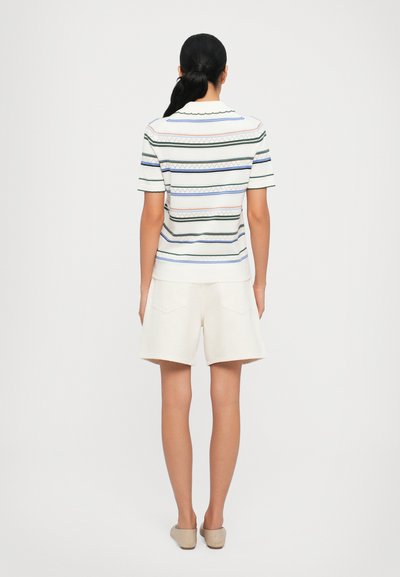 Top a maniche corte a righe bianche, con righe orizzontali verdi, blu e rosa, e colletto a scollatura ondulata. Abbinato a pantaloni corti color crema.