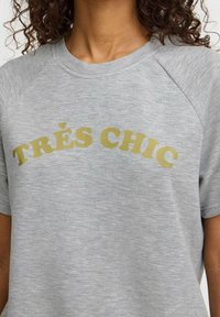 Grå kortärmad t-shirt med rundad "TRÈS CHIC" i fet gulddetalj. Mjuk, texturerad tyg med en avslappnad passform och raglanärmar.